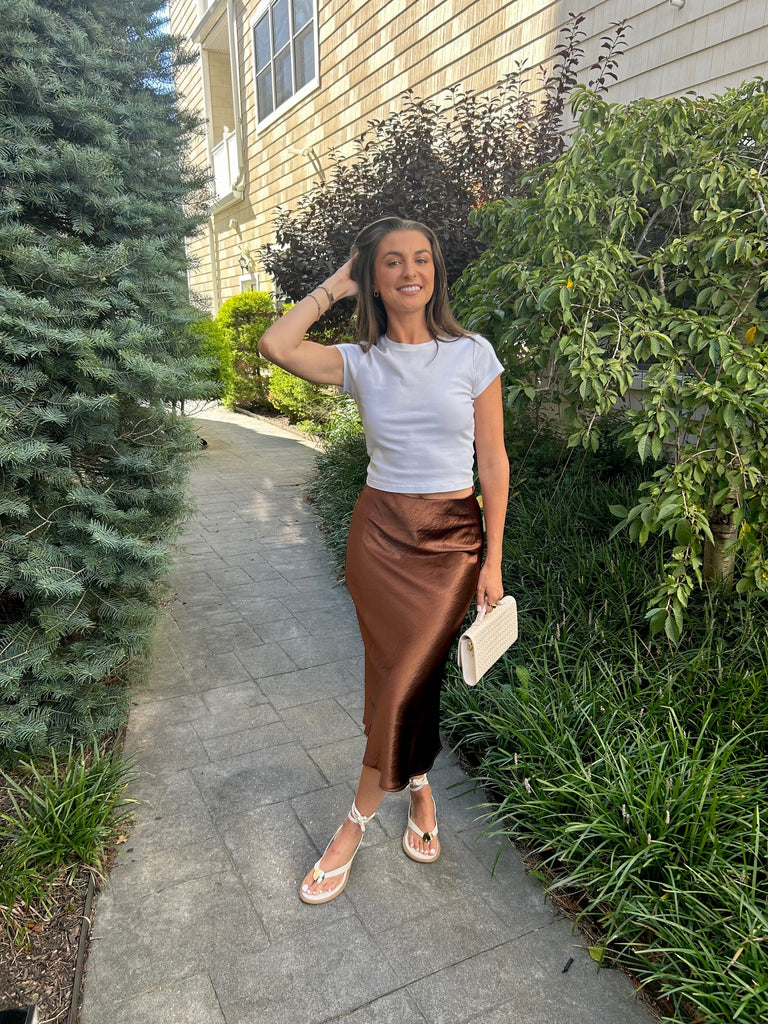 Woman in a white top and brown skirt standing on a sidewalk with greenery around.