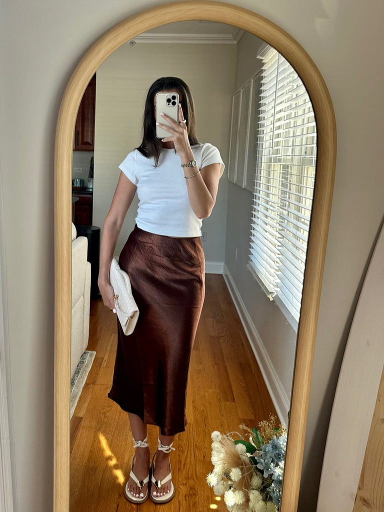 Person taking a mirror selfie wearing a white top and brown skirt in a room with wooden flooring.