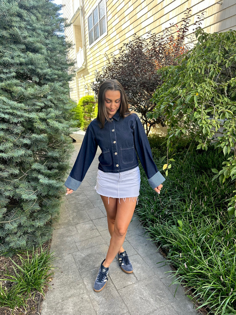 Woman in a denim jacket and white skirt walking on a sidewalk with greenery around.